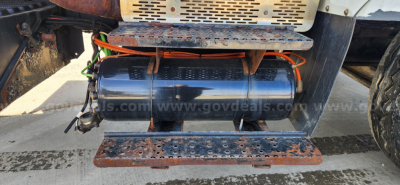 Truck undercarriage showing cylindrical fuel or air tank with orange wiring and metal steps; diesel engine bay components visible.