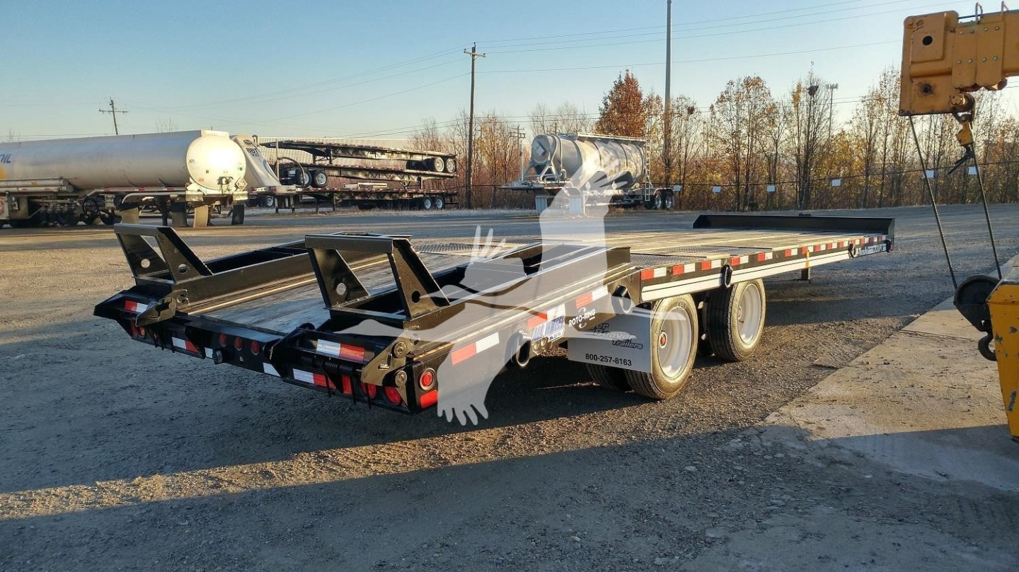 Trailer deck with multiple axles showing long flatbed with side rails and rear ramps. on-site lot with other equipment in background.