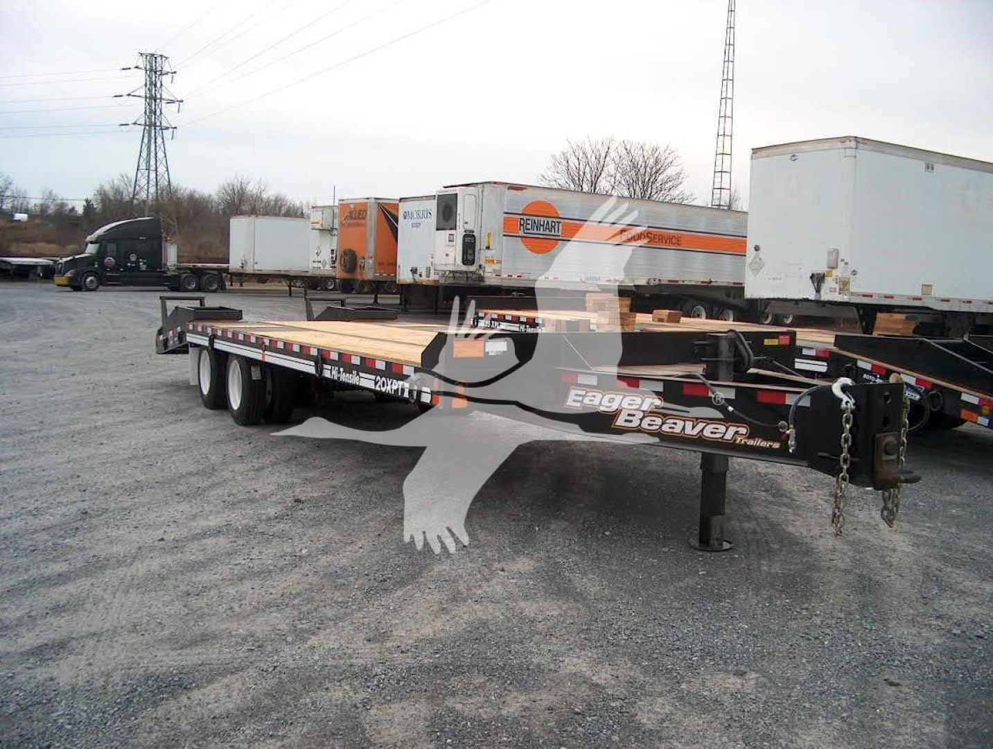 Flatbed trailer showing 5-axle beavertail with Eager Beaver branding and orange/black color scheme, hitch and chains visible in foreground.