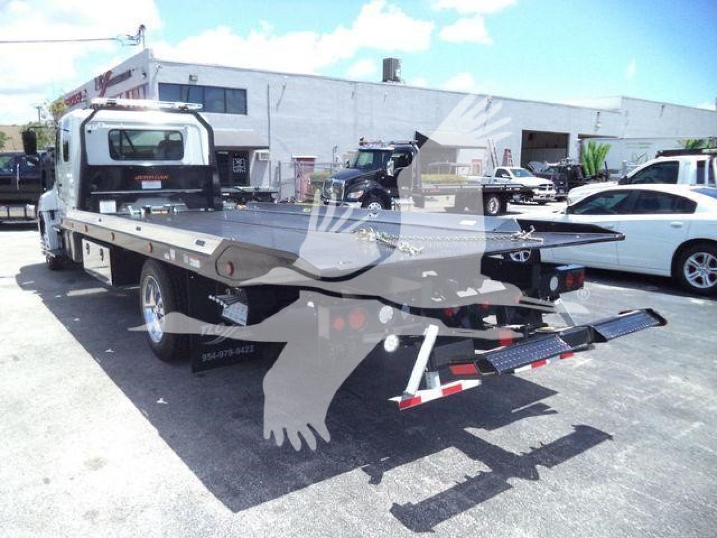 Flatbed rollback tow truck in lot with rear ramps deployed and Hino L6 chassis visible.