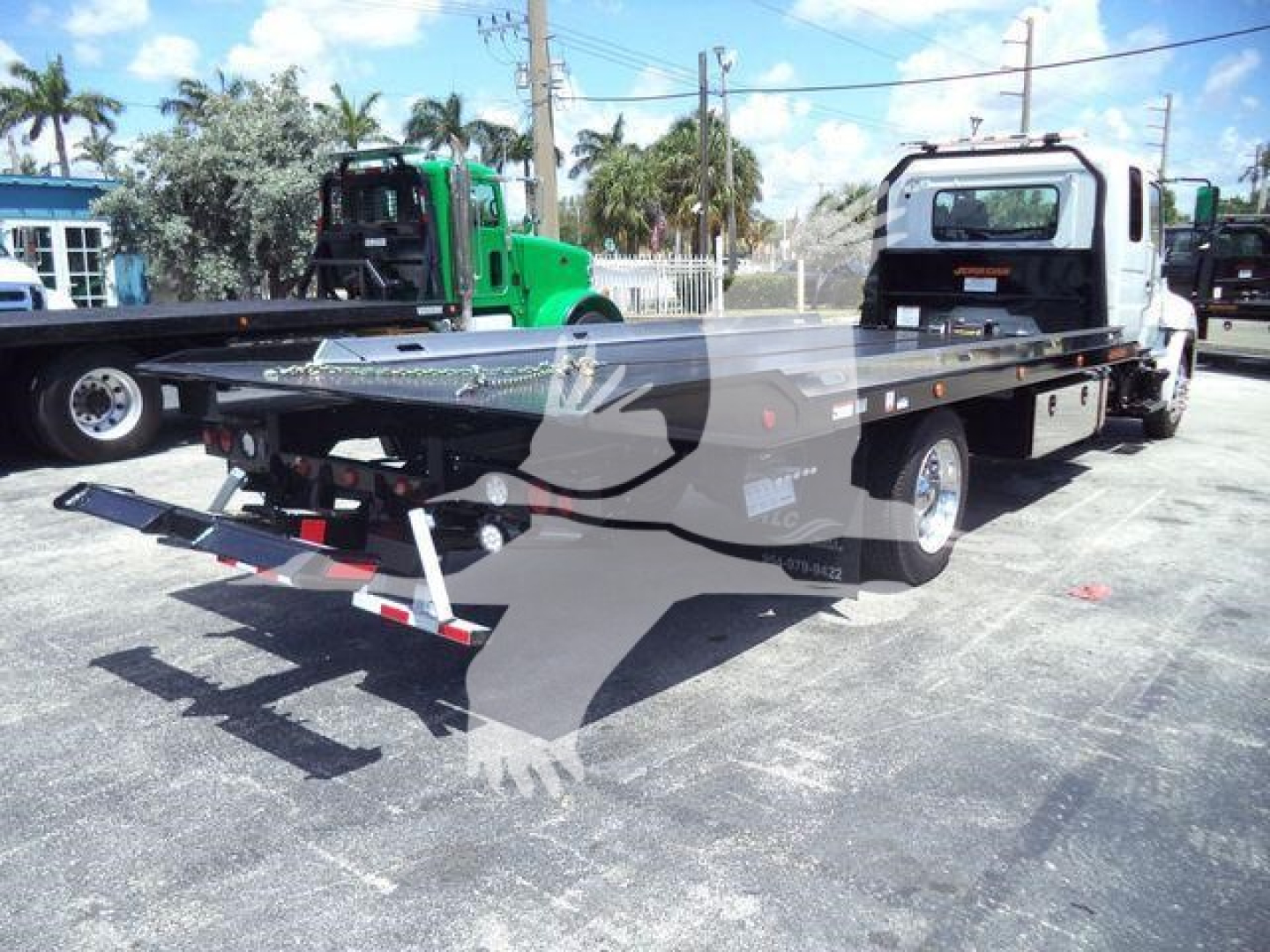 Flatbed tow truck with a white cab and long black flatbed, rollback equipment, and a wheel-lift setup visible.