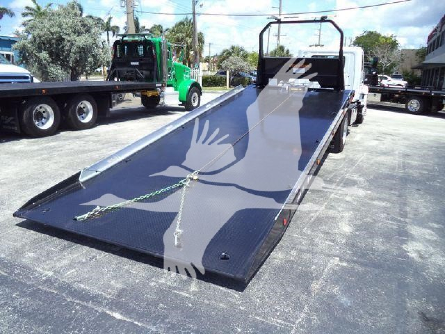 Flatbed rollback truck with ramp down showing black platform and metal frame, cab area in background, no visible branding text.