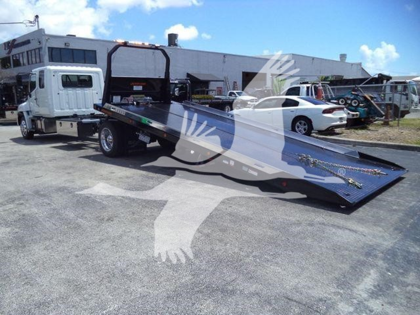Truck with flatbed rollback loader in a lot. White cab connected to a large black flatbed with ramp, other vehicles and a warehouse in background.