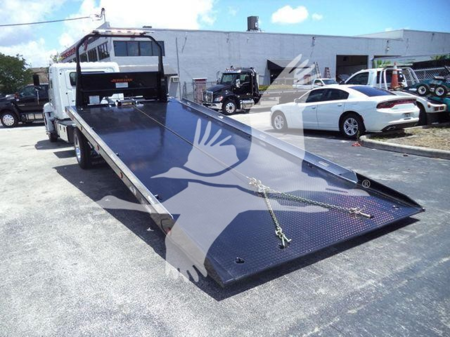 2026 Hino L6 rollback tow truck with flatbed carrying surface visible from rear angle; black rubber-coated deck, front cab, hydraulic system area at rear left.