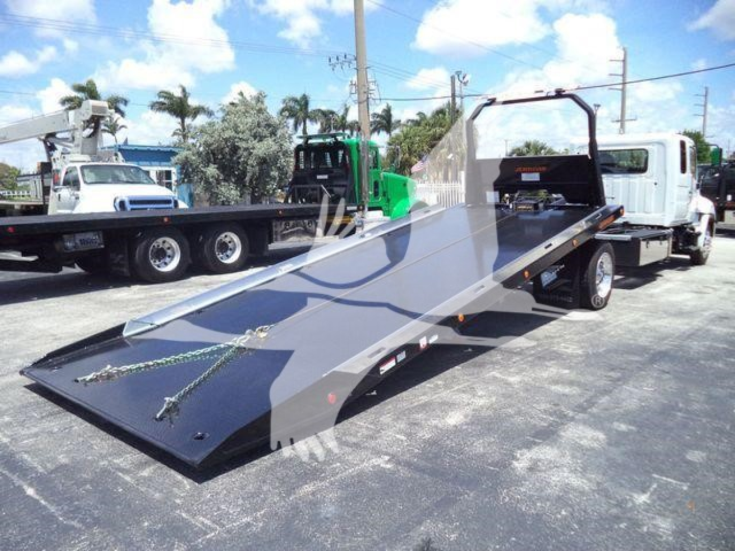 Truck flatbed rollback tilt bed showing metal platform with winches and safety chains, white cab behind, in outdoor lot.