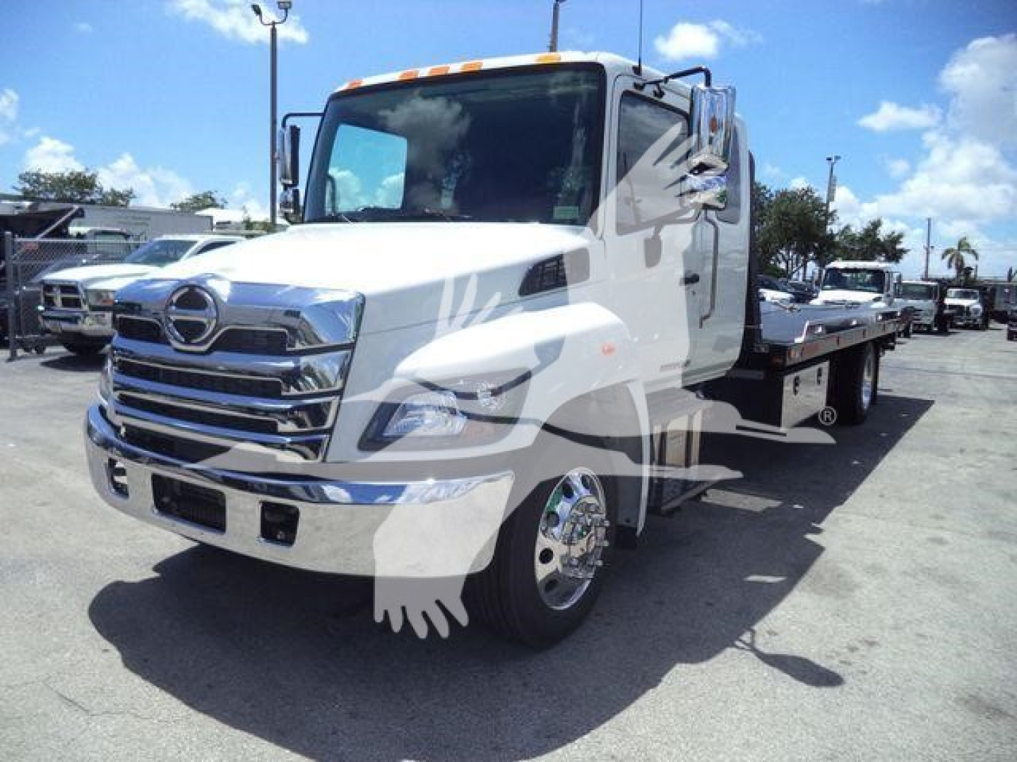 Truck engine bay view showing Hino L6 cab and a flatbed rollback bed, front chrome grille, headlights, and side mirrors.