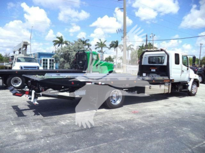 Hino L6 rollback truck with a flatbed tow bed and white cab. Visible in the background are other trucks and a blue building.