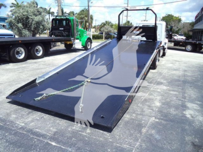 Flatbed rollback truck with ramp down showing black platform and metal frame, cab area in background, no visible branding text.