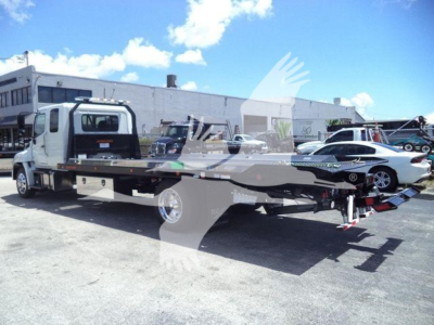 Flatbed tow truck with a white cab and grey flatbed carrying a car; blue sky with clouds; dealership lot in background.