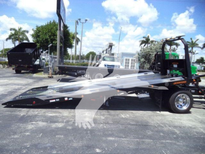 Flatbed tow truck in a dealership lot showing a Hino L6 rollback bed and equipment; truck with ramped platform, wheel chocks, and branding faint on bed.