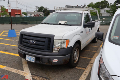 Ford F-150 pickup truck with utility bed and ladder rack, front bumper with stickers, license plate visible, in a parking lot.