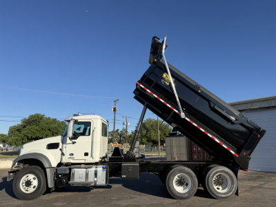 Truck dump body in raised position on a white Mack GR64F cabover chassis, three rear axles, black dumped bed with safety supports visible.