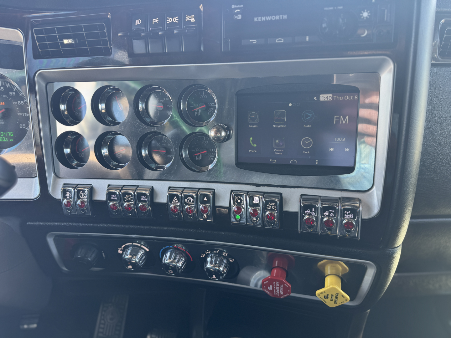 Kenworth W900L dash panel showing multiple gauges and a touchscreen display with various switches and colored knobs.