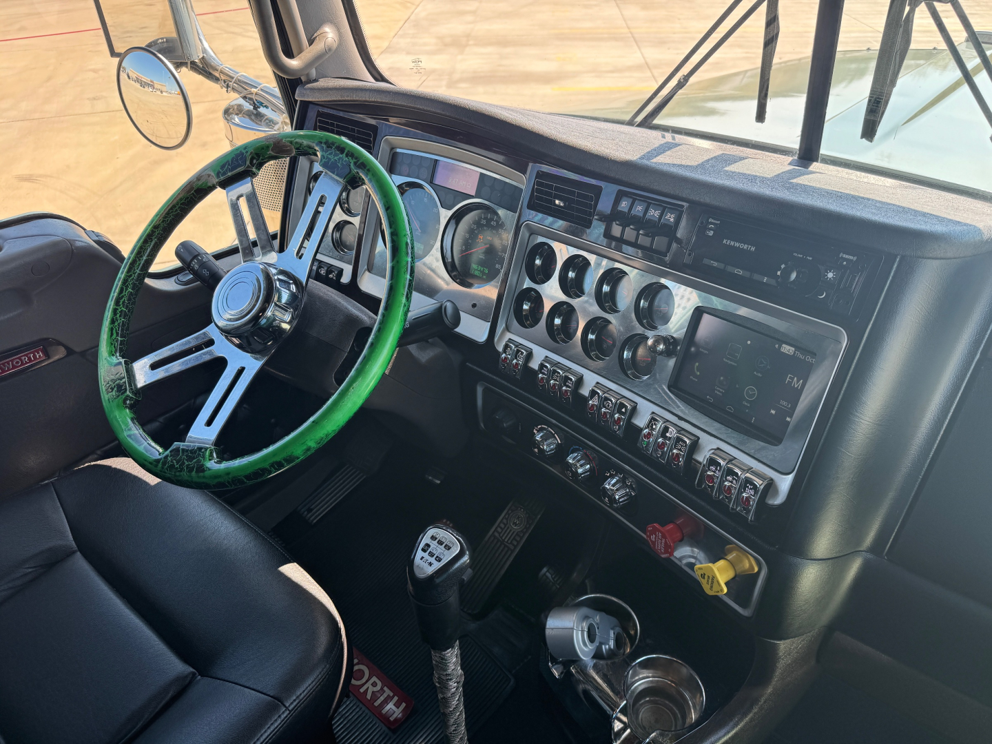 Kenworth W900L interior dashboard view showing steering wheel with green cover, multi-gauge cluster, and row of switches.