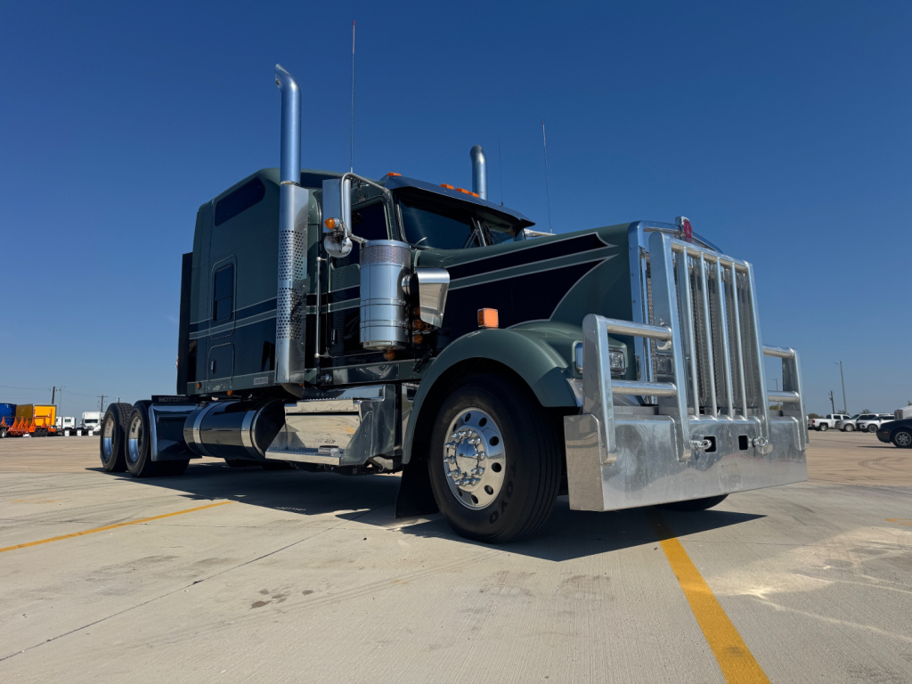 Kenworth W900L conventional sleeper truck in a parking lot, side view showing chrome exhaust stacks, large grille, dual rear axles, and polished metal accents.