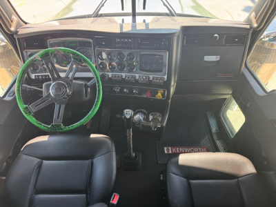 Kenworth W900L cab interior entry showing black steering wheel with green cover, dashboard with gauges, three stalks gear lever and manual shifter; Kenworth badge on floor mat visible.