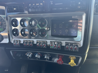 Kenworth W900L dash panel showing multiple gauges and a touchscreen display with various switches and colored knobs.