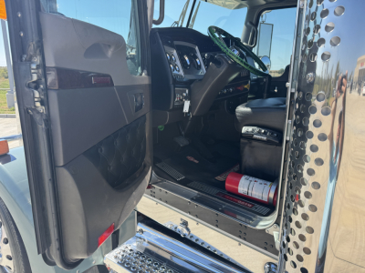 Kenworth W900L cab interior view with open driver door showing steering wheel, dashboard, and entry step; chrome exterior panels reflected; warning labels near door sill.