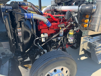 Kenworth W900L engine bay view showing Cummins X15 engine, radiator, hoses, and front wheel. Visible components include coolant reservoir, turbo piping, and air intake.