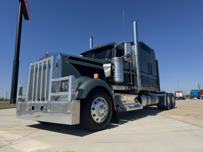 Kenworth W900L conventional sleeper truck in blue-gray with chrome grille guard and dual exhaust stacks, large front bumper, side fueling tank, and chrome wheels.