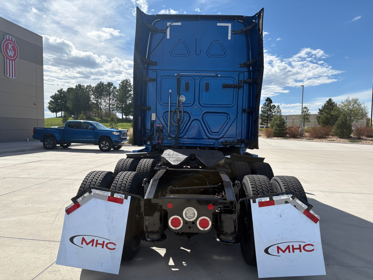 Truck rear view of 2022 Freightliner PT126064ST tractor showing dual rear axles, mud flaps with MHC logo, and blue sleeper cab; marketplace lot in background.