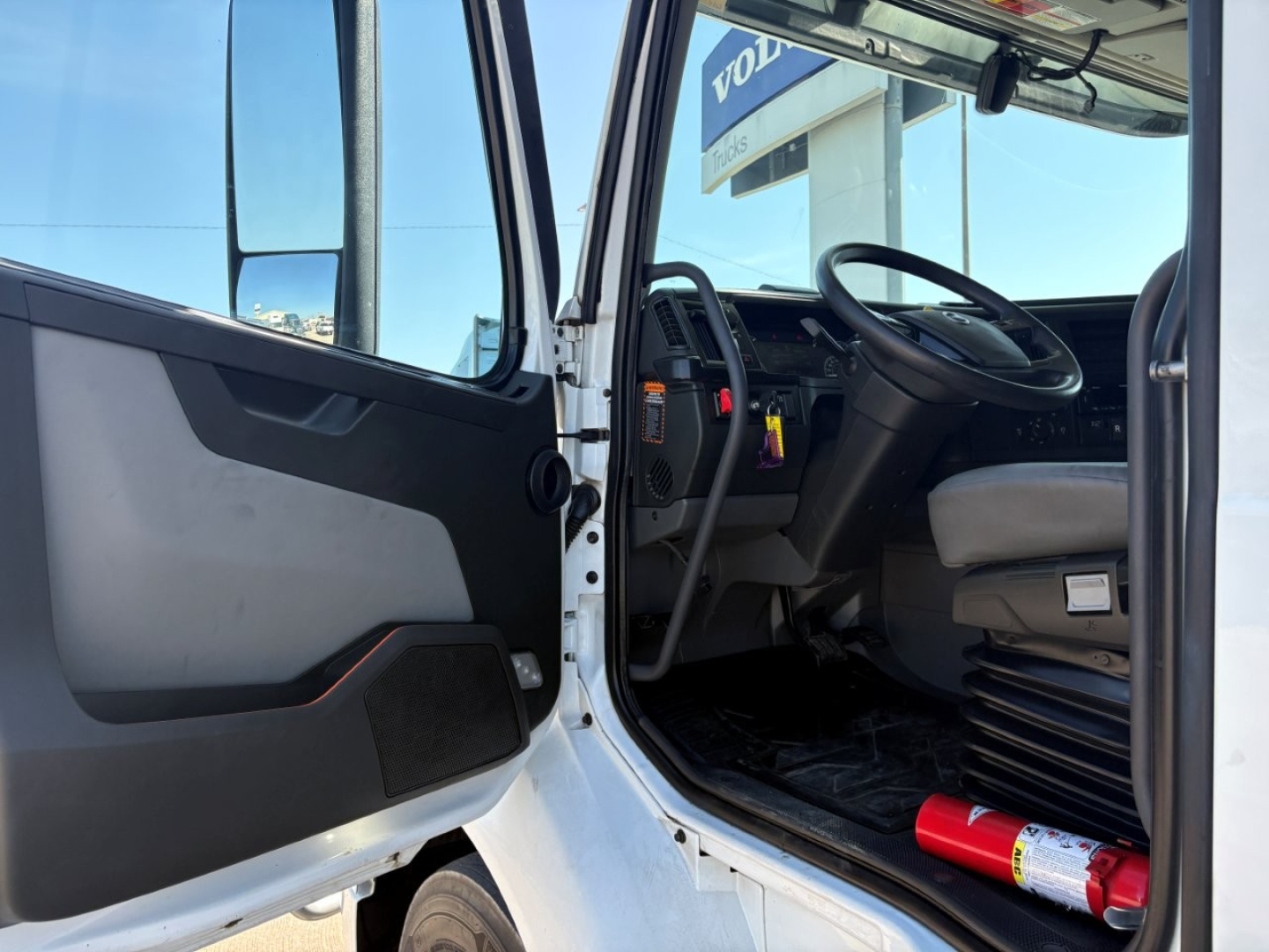 Volvo VNR64T660 truck cab interior view with open driver door showing steering wheel and dashboard; seat, pedals, and fire extinguisher on floor; exterior white paint and window.