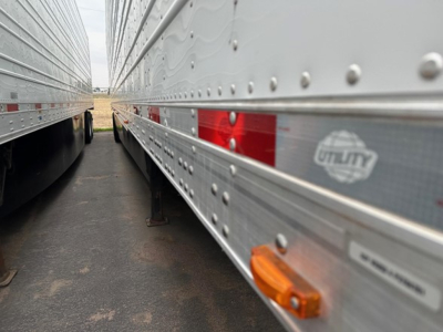 Two parallel semi-trailer sides in a lot, with reflective metal siding and small orange side marker light visible; brand emblem and riveted panels present along the length.
