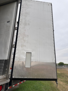 Trailer side door and lower rear corner of a 2023 Utility 3000R reefer trailer showing smooth aluminum exterior with riveted edge and a small rectangular access panel.