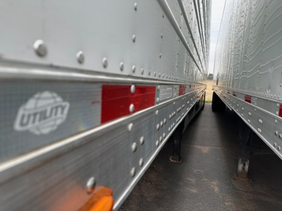 Reefer trailer side view showing Utility logo and riveted aluminum walls between two trailers; wheel chocks visible on ground; red reflective panels.