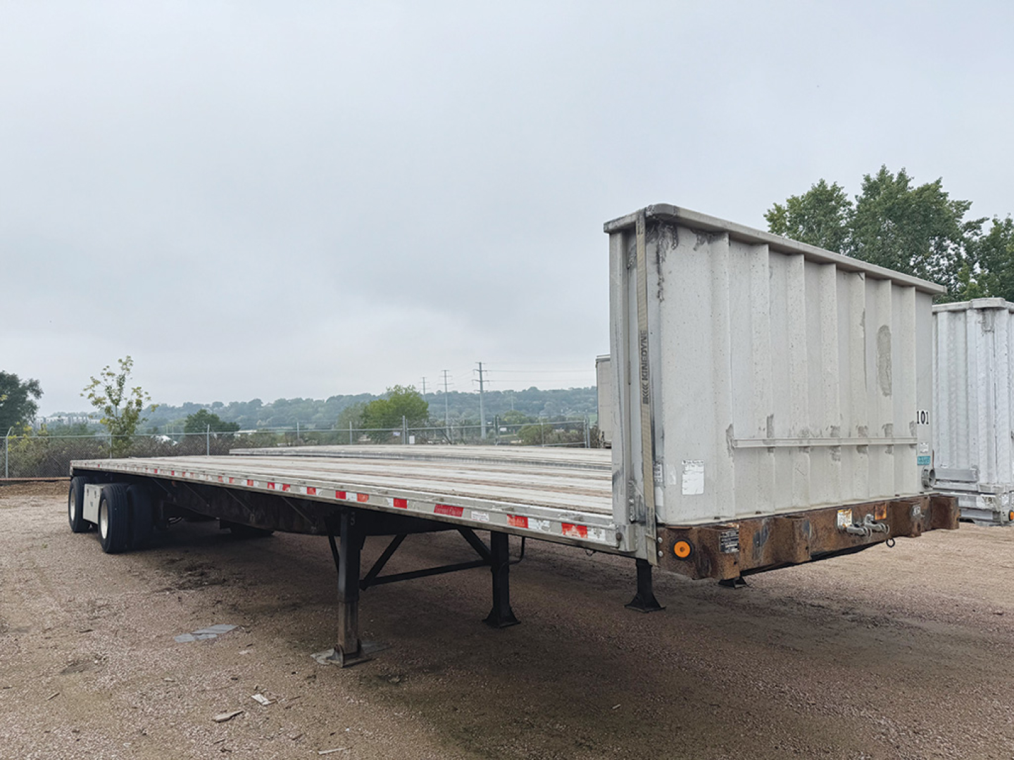 Flatbed trailer showing long aluminum and steel deck with elevated gooseneck style end and rear tires visible.