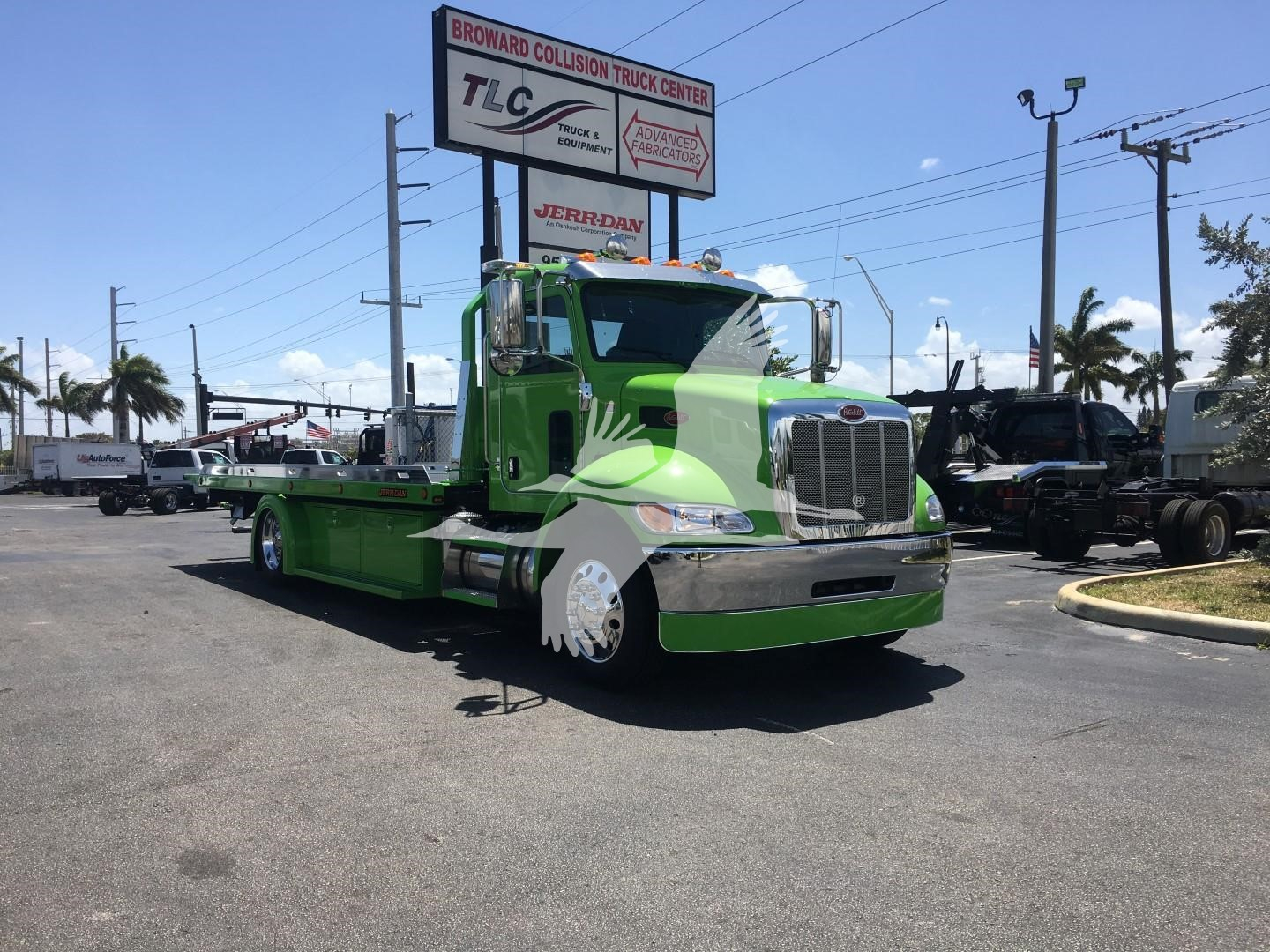 Green 2022 Peterbilt 337 rollback truck with front chrome grille and long flatbed, parked at a dealership lot with signboards in background.