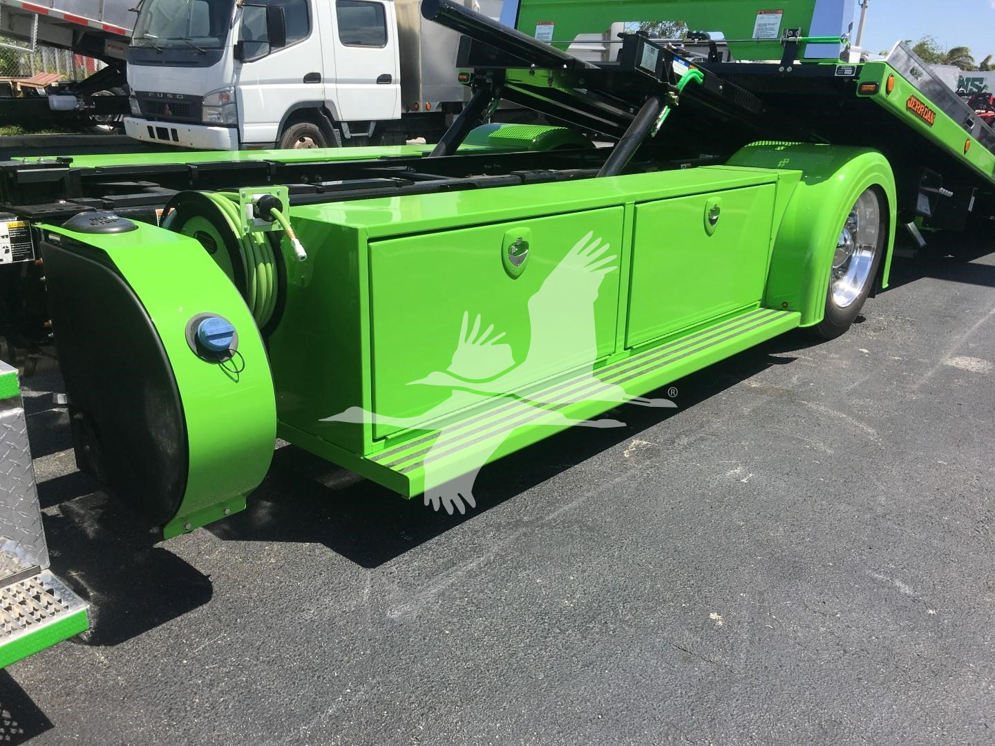 Green Peterbilt rollback tow truck side view showing storage compartments and wheel cover. Visible text: none readable.