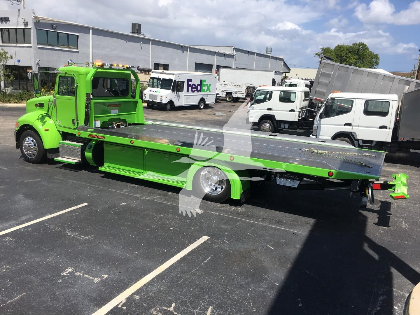 Green Peterbilt 337 rollback tow truck side view showing long aluminum flatbed and wheel-lift, with FedEx truck in background.
