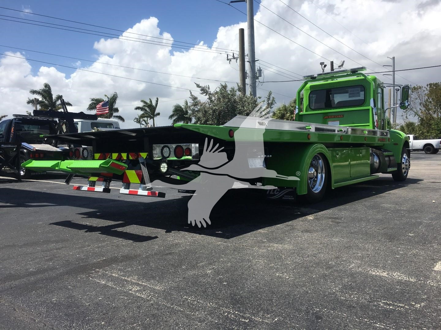 Green Peterbilt 337 rollback tow truck from rear-left angle showing flatbed, dual rear axles, and branding. No legible serials or text visible on side; background includes other trucks and palm trees.