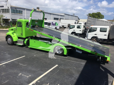 Bright green 2022 Peterbilt 337 rollback tow truck with a tilted flatbed in a dealership lot, FedEx and other trucks in background.