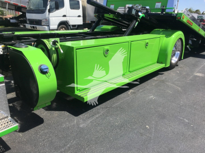 Green Peterbilt rollback tow truck side view showing storage compartments and wheel cover. Visible text: none readable.