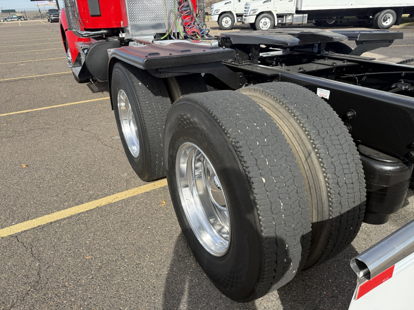 Kenworth T880 chassis showing dual rear axles and tires, with fuel tanks and frame visible; red cab in background with parking lot”.
