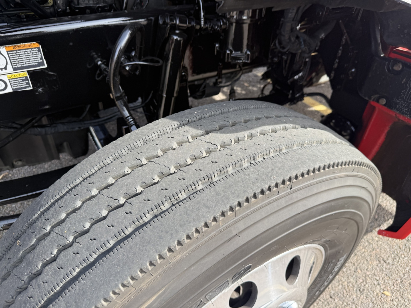 Kenworth T880 wheel and front suspension area showing tire treads and rim, with portions of the truck frame and hydraulic lines in view.