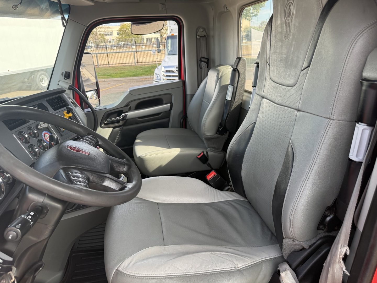 Kenworth T880 truck interior showing driver seat and passenger seat. Steering wheel with dashboard controls, gray fabric seats, door panel with handles, seat belts; windows reveal outdoor area.