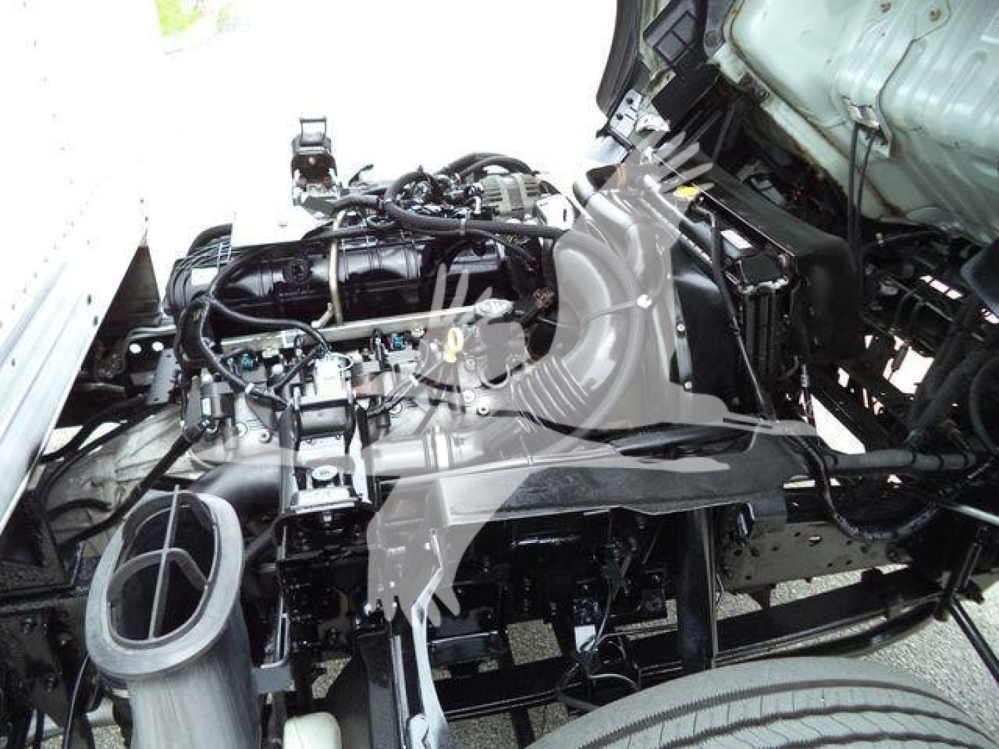 Isuzu NPR HD engine bay showing exposed engine components and intake manifold with surrounding chassis. View focuses on front engine area, hoses, and mounting brackets.