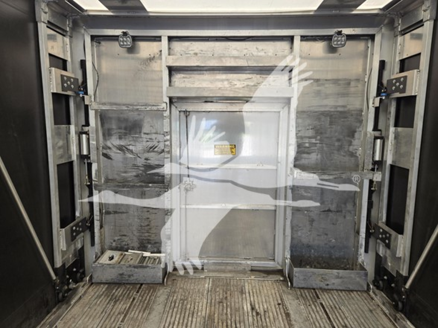 Trailer interior showing aluminum flatbed trailer with sidewalls and rear ramp area; metallic surfaces, tiedown rails, and doorway hatch in center. 