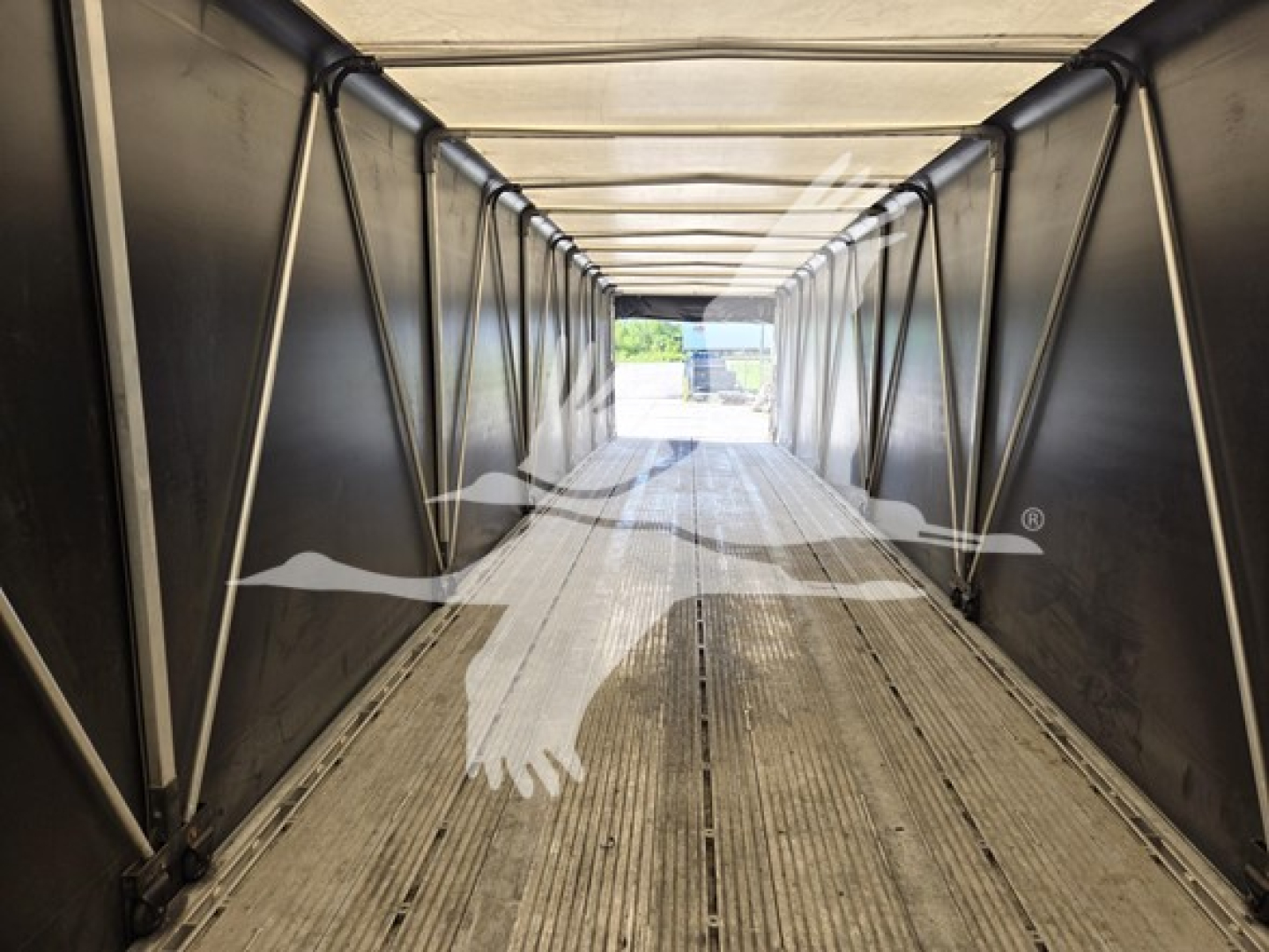 Aluminum flatbed trailer interior with ribbed floor and side rails, long corridor viewpoint toward open end.