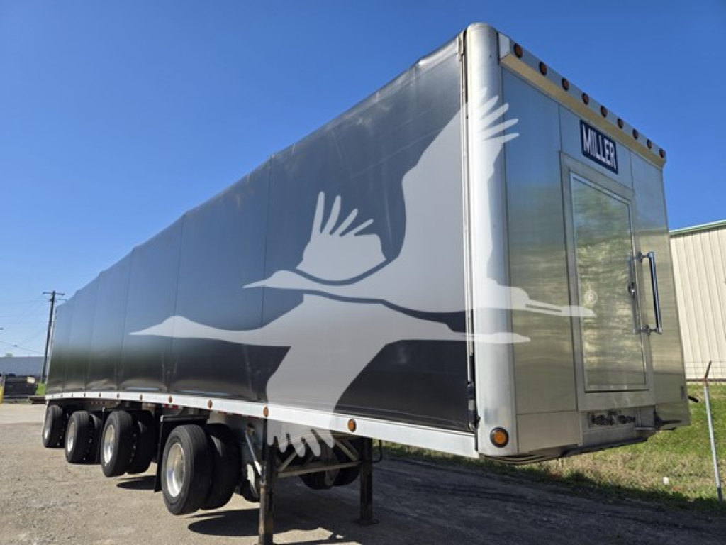 Trailer side view showing large white bird graphic on aluminum flatbed trailer with Miller logo at rear and triple-axle configuration.