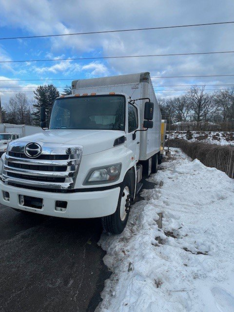 White Hino 268 box truck in snow, side view showing cab and box with riveted doors and reflective lights on top. No legible labels or numbers visible on the vehicle itself.