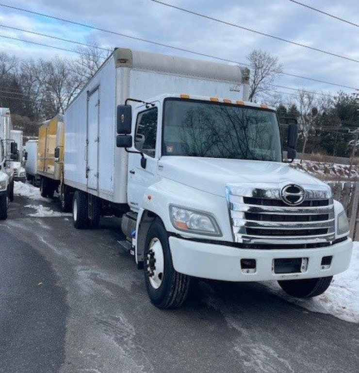 Truck: 2016 Hino 268 box van with white cab and dual rear wheels, front grille with chrome bars, large side mirrors, cargo box attached.