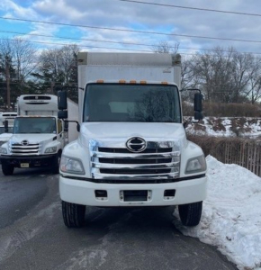 Front view of a white 2016 Hino 268 box van truck showing chrome grille, Hino emblem, large bumper, and snow on roadside.