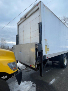 Truck rear of a box van with liftgate and yellow cab visible on left; VIN/sticker area on box side.