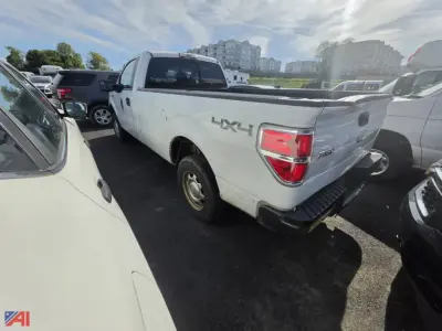 2012 Ford F-150 pickup truck in a lot, side/rear view showing 4x4 badge on the rear quarter, white exterior, tail lights, bed cap