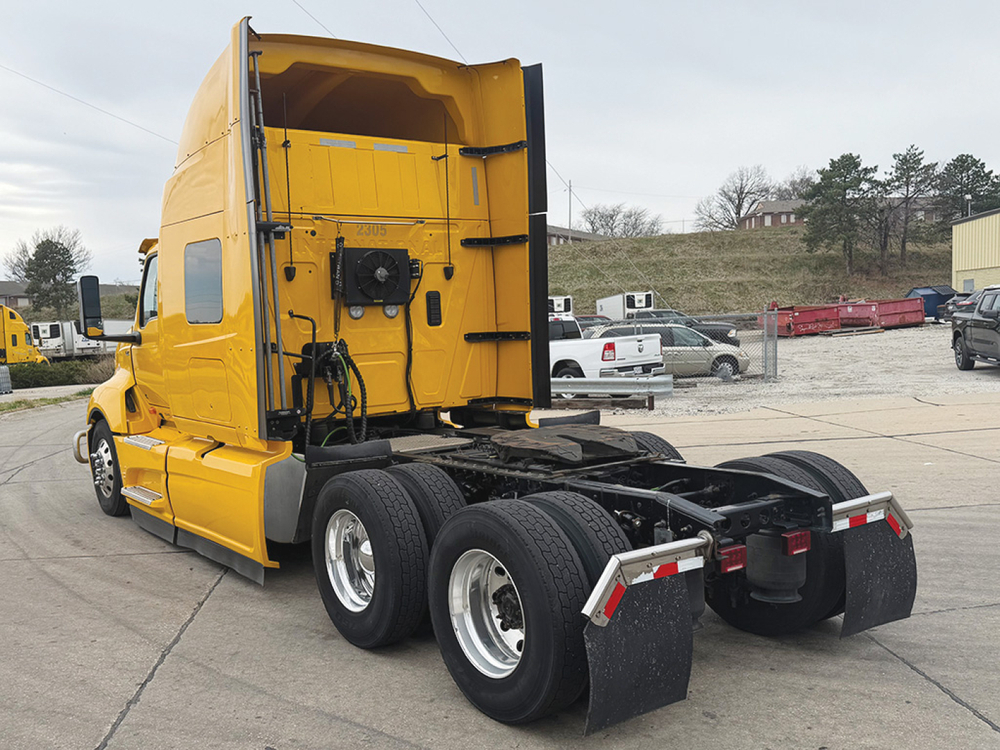 Truck tractor in yellow showing rear/side view of the fifth wheel and dual rear axles with mud flaps. Text not visible; image focuses on cab and chassis.