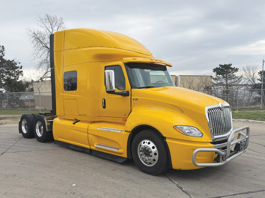 Large yellow International LT625 semi-truck, sleeper cab, front grille guard, side steps, chrome wheels, cargo trailer hitch, in a parking lot.
